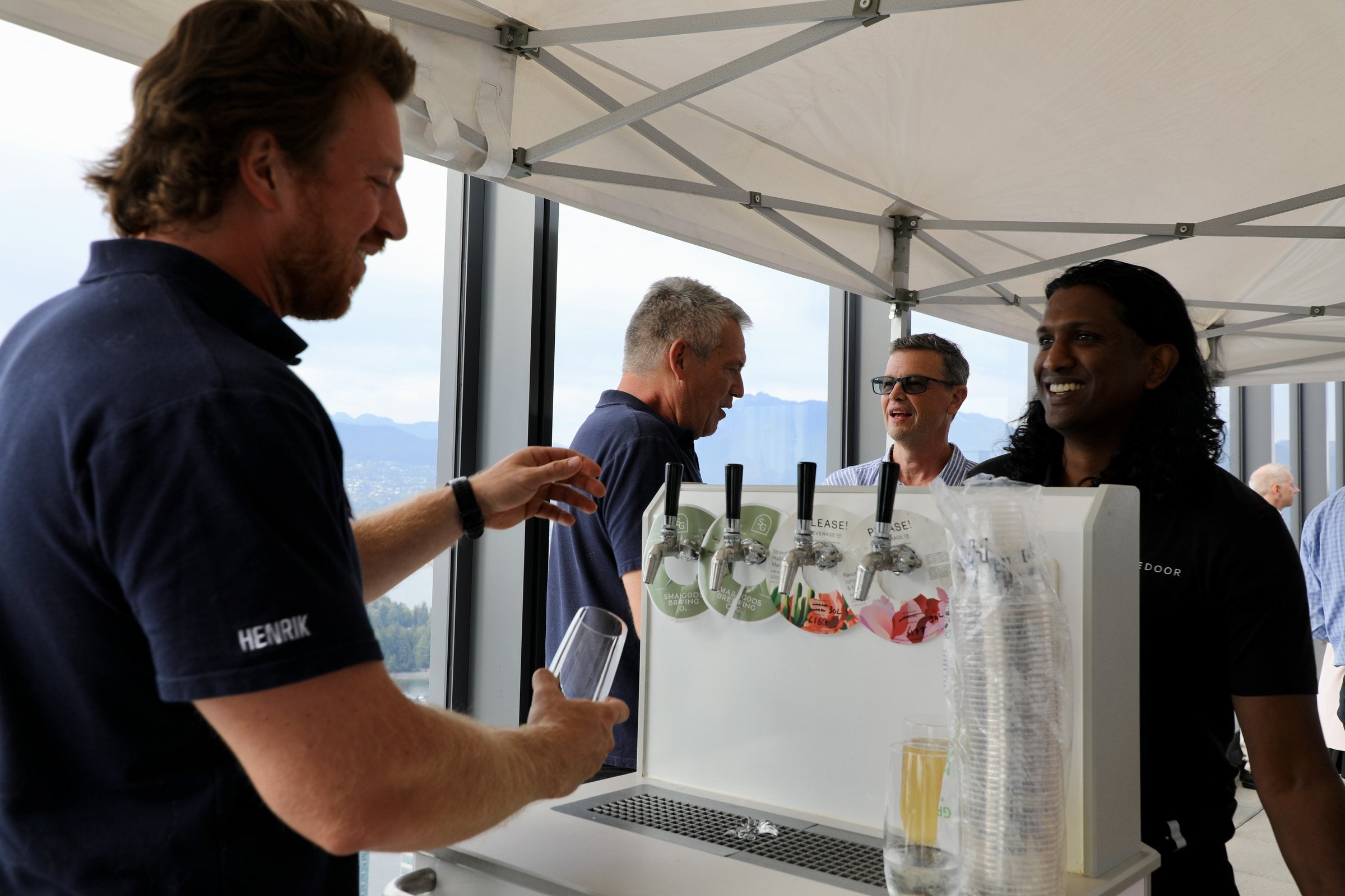 A Henrik team member handing out a glass at a four-tap draft bar under a tent with a view over Vancouver